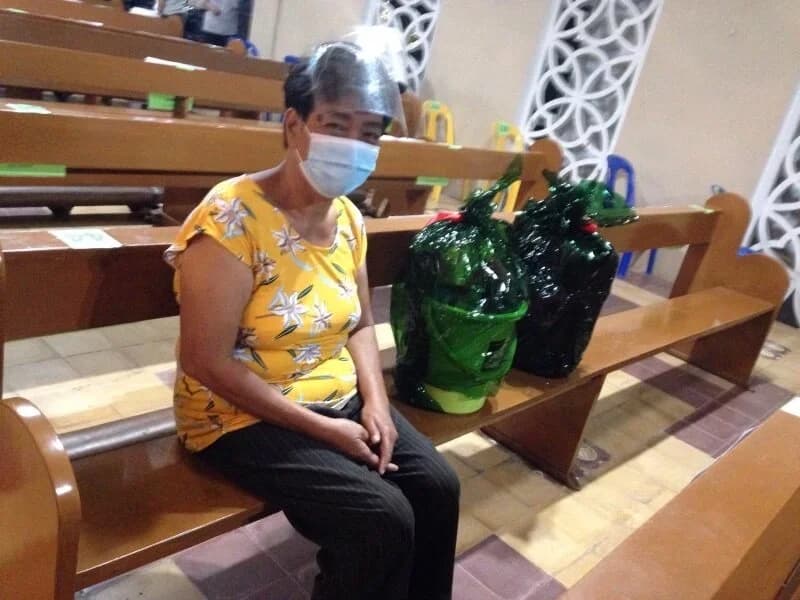 A masked woman sits alone on a wooden church pew with two green-wrapped baskets tied with red ribbon on the bench beside her.
