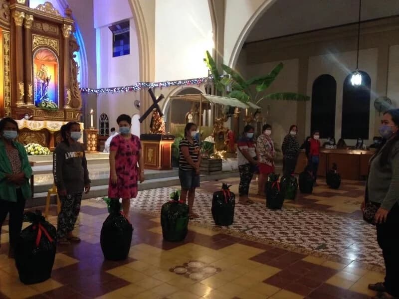 People stand well apart across the tiled floor of a church, the altar lit behind them, a wrapped basket on the floor at each person's feet.
