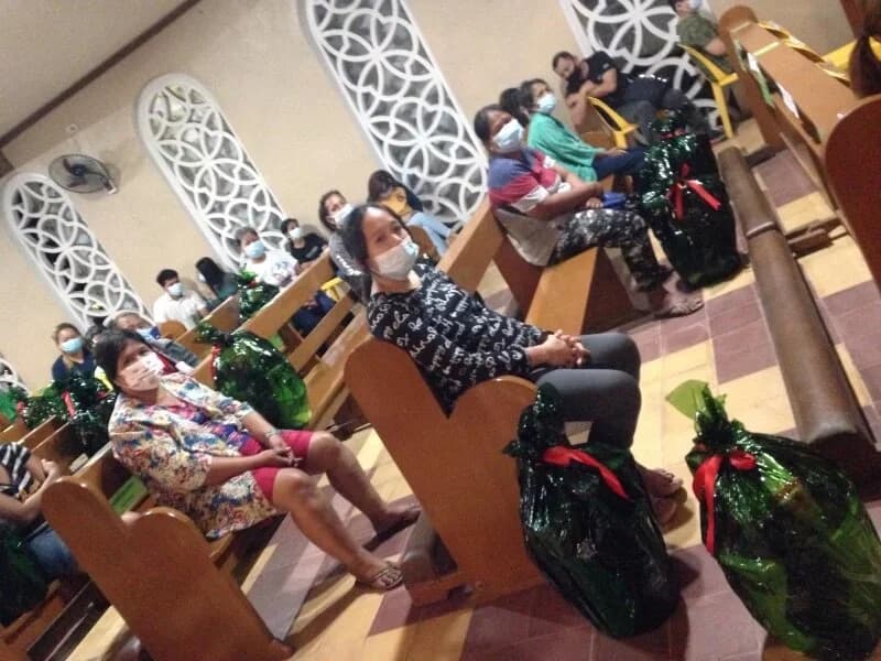 Masked people sit spaced along the church pews, each with green-wrapped baskets tied with red ribbon on the floor beside them.