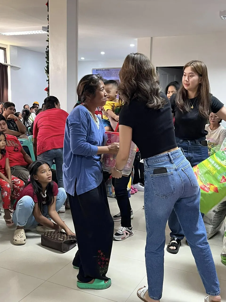 A volunteer in a black shirt hands goods to a woman in a blue shirt inside the hall, with families and young children seated on chairs behind them.