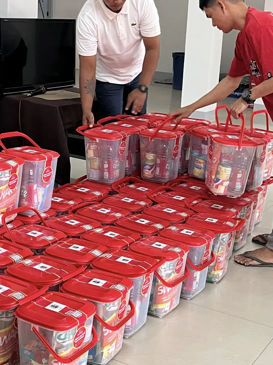 Two volunteers lift red pails by their handles; the floor around them is covered with pails packed with instant noodles, coffee and canned goods.