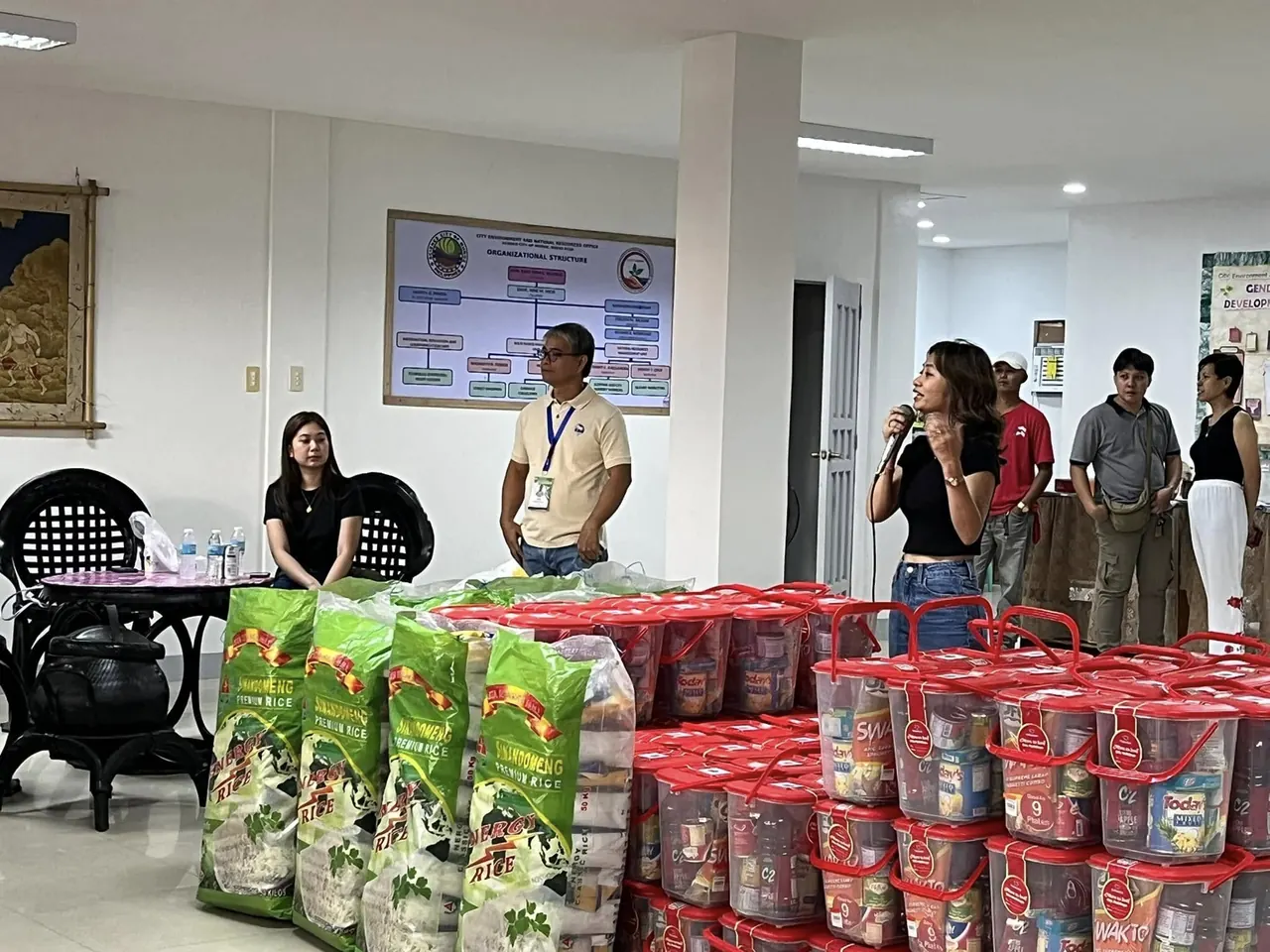 Sacks of rice stacked beside rows of red grocery pails on a hall floor; a woman speaks into a microphone while officials sit at a table beneath the office's organisational chart.