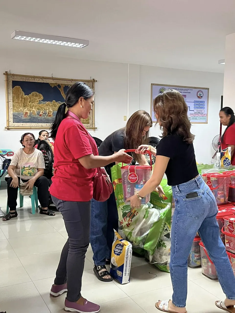 A volunteer in a black shirt passes a bag of produce and a sack of rice to a woman in a red shirt, with recipients seated along the wall behind.