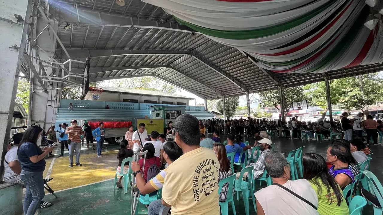 A covered basketball court draped with bunting, a teal truck stacked with red bags parked inside it, and rows of people seated on plastic chairs waiting.