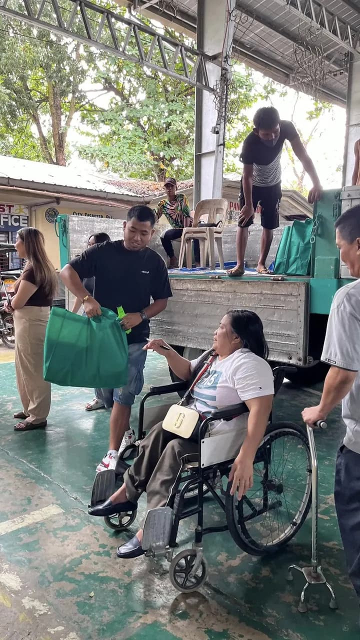 A woman in a wheelchair is handed a green eco bag by a volunteer standing at the tailgate of the teal truck.