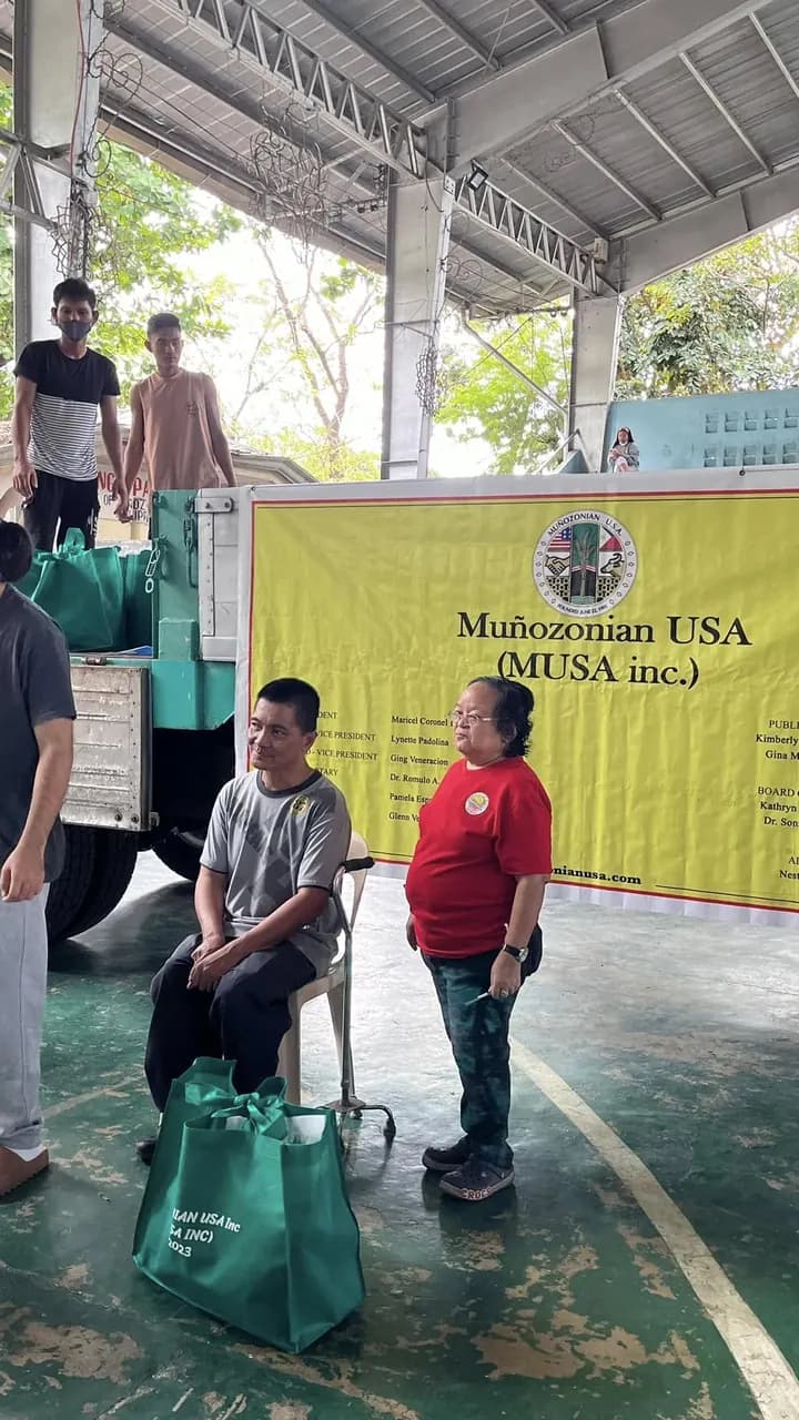 A man sits on a plastic chair with a walking cane beside him and a green eco bag on the court floor; a woman in a red Muñozonian USA shirt stands next to him in front of the yellow MUSA tarpaulin.