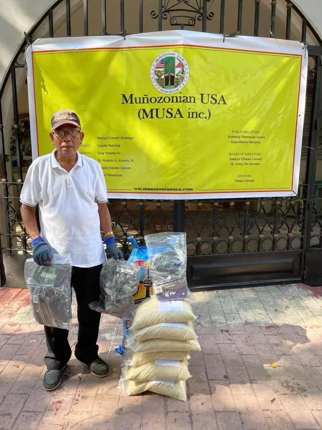 A man in a white shirt and cap stands in front of the yellow Muñozonian USA tarpaulin at a church gate, holding a wrapped packet, with bags of rice on the pavement at his feet.