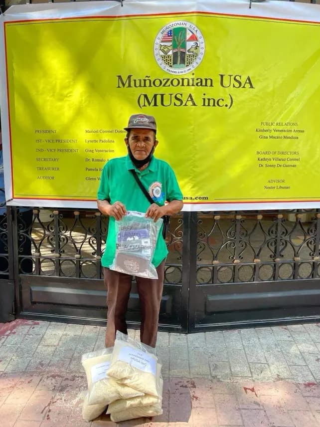 A woman in a green shirt and cap holds up a clear packet in front of the MUSA tarpaulin, bags of rice on the ground in front of her.