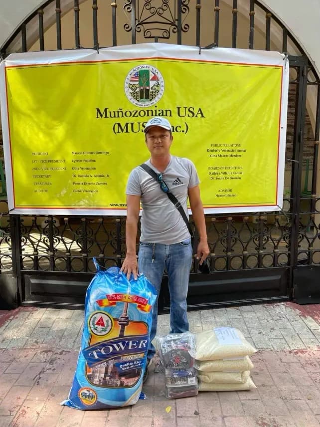A man with a shoulder bag stands by the tarpaulin beside a blue branded sack of rice, a packet of goods and further bags of rice at his feet.