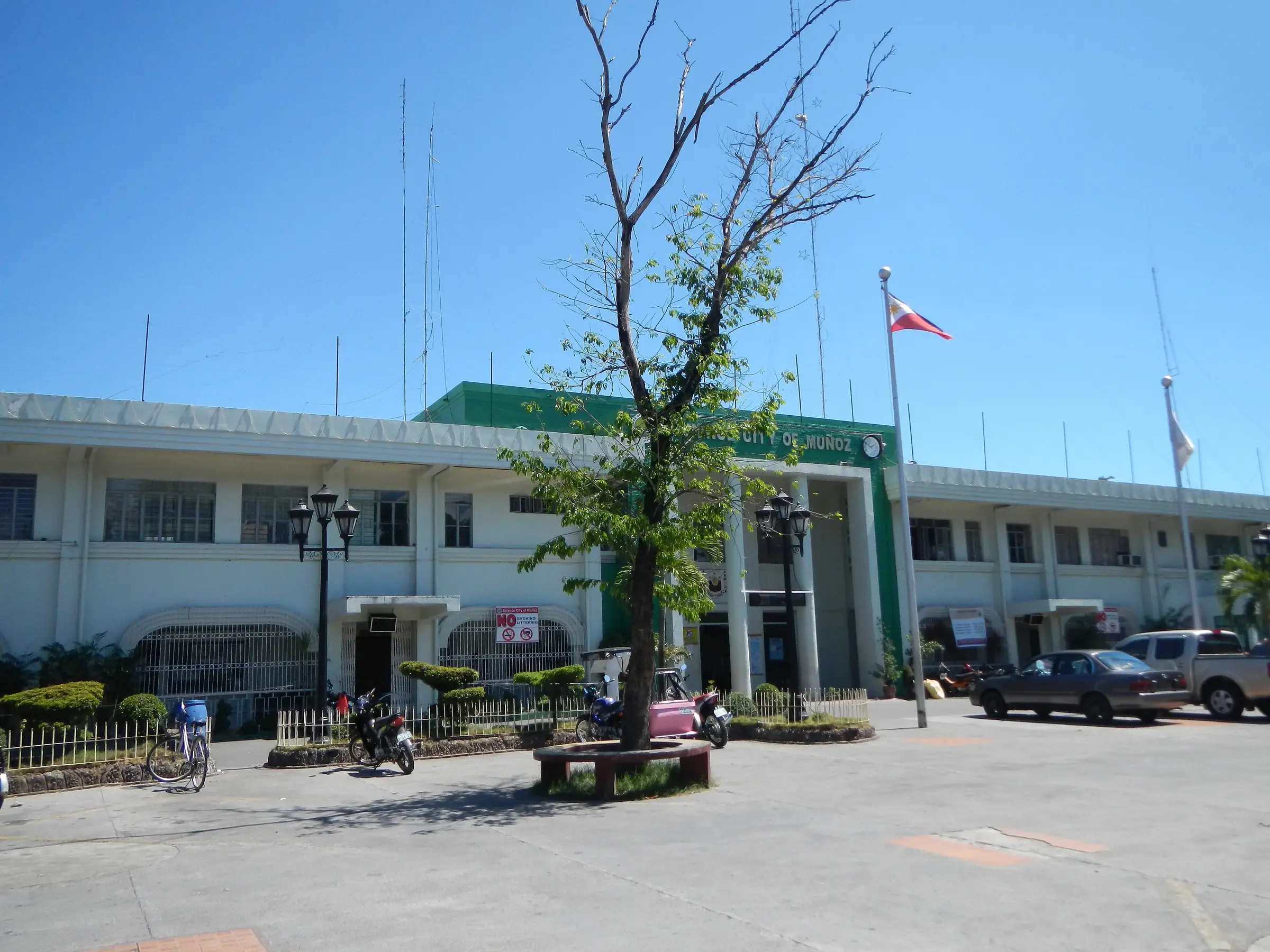The long two-storey city hall of Muñoz with a green-roofed entrance, a bare tree and a Philippine flag in the plaza before it.